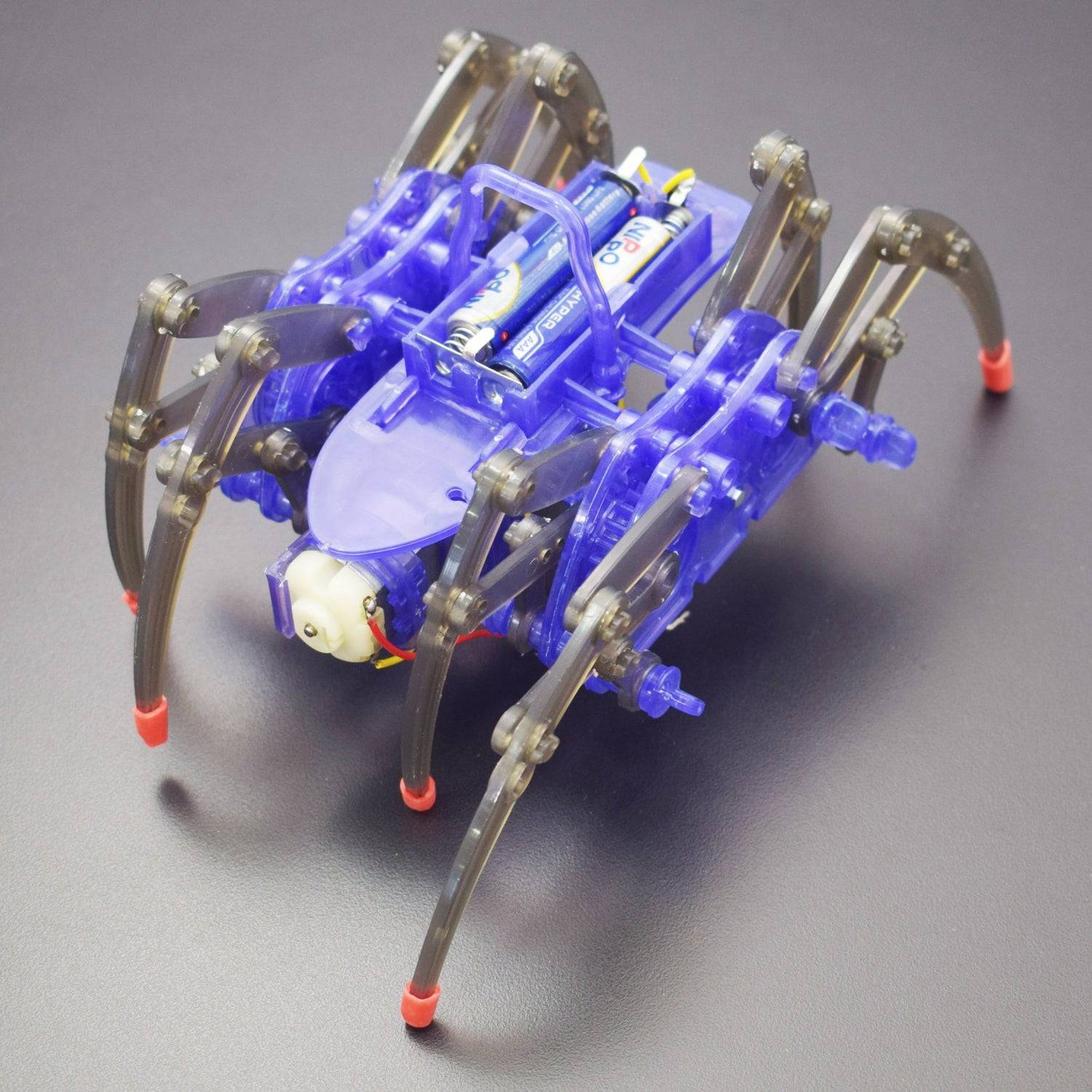 A blue, transparent electric spider with eight legs and visible battery compartments stands on a dark surface, showcasing its mechanical design and translucent components.