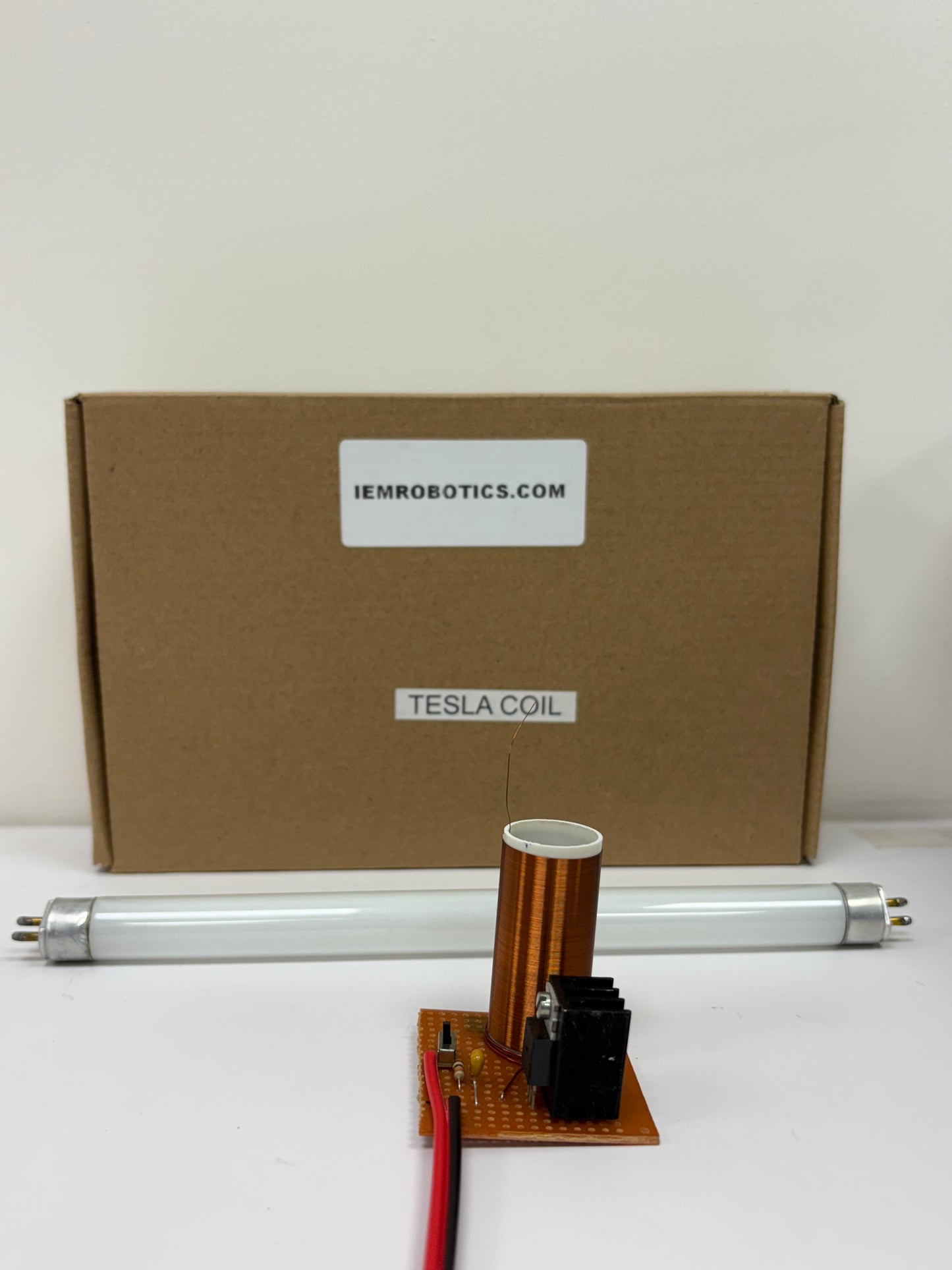 A small Tesla Coil Science Kit with a cylindrical coil on a circuit board sits in front of a cardboard box labeled iemrobotics.com and Tesla Coil. A fluorescent tube rests horizontally beside the coil, offering hands-on learning opportunities.